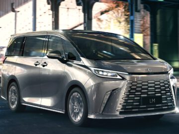Een zilverkleurige bestelwagen met de modelnaam "Lexus LM" op de voorste kentekenplaat rijdt op een weg onder een brug door.