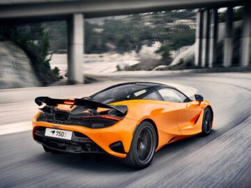 De McLaren 750S, een oranje sportwagen met aerodynamisch ontwerp, snelt over een bochtige weg onder een brug door.