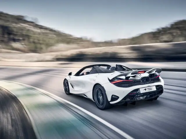 Een witte McLaren 750S met verhoogde achtervleugel rijdt met hoge snelheid over een bochtige weg. Bewegingsonscherpte benadrukt de snelheid van de auto en de golvende landschapsachtergrond.