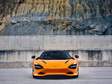 Een oranje McLaren 750S-sportwagen staat geparkeerd voor een betonnen barrière, met een rotsachtige klif op de achtergrond.