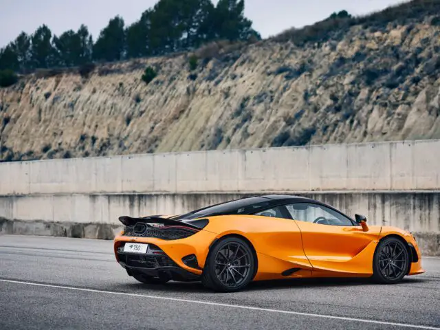 Een feloranje McLaren 750S-sportwagen staat geparkeerd op een weg met een betonnen barrière en een heuvel op de achtergrond.