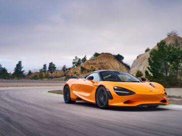 Een feloranje McLaren 750S-sportwagen rijdt op een gebogen weg met heuvels en bomen op de achtergrond onder een bewolkte hemel.