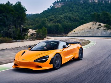 Een knaloranje McLaren 750S rijdt op een bochtige bergweg, omgeven door beboste heuvels.