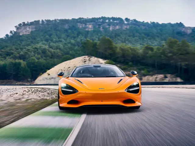 Een oranje McLaren 750S snelt over een verharde weg, omlijst door een prachtige achtergrond van bomen en bergen.