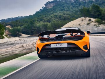 Oranje sportwagen met zichtbaar kenteken "750S" rijdend op een verharde weg omgeven door bomen en heuvels. Het zicht is vanaf de achterkant en laat het strakke ontwerp van de McLaren 750S zien.