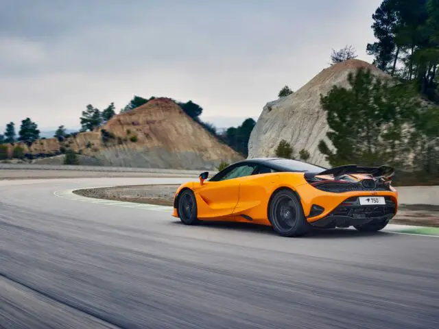 We zien een levendige oranje McLaren 750S-sportwagen rijden op een bochtige weg met rotsachtige heuvels en bomen op de achtergrond.