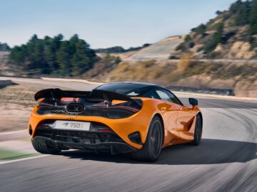 Een feloranje McLaren 750S-sportwagen rijdt op een bochtige weg. De auto heeft een "750S" -badge op de achterkant en een zichtbare achterspoiler. Bomen en heuvels bevinden zich op de achtergrond onder een heldere hemel.