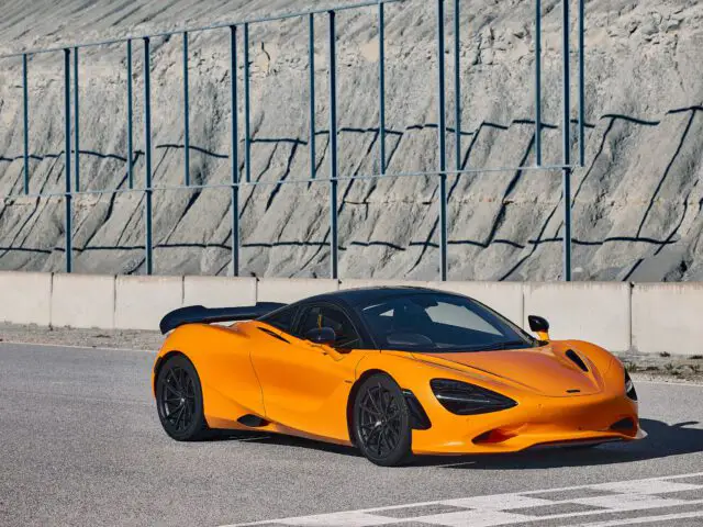 Een feloranje McLaren 750S-sportwagen staat geparkeerd op een verhard terrein nabij een rotswand met metalen barrières.