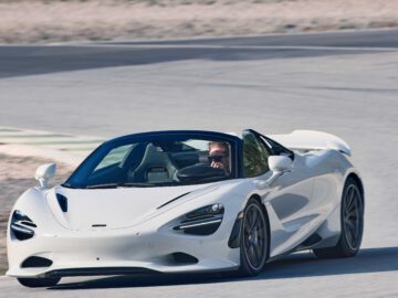 Een witte McLaren 750S cabrioletsportwagen rijdt over een verharde weg met een persoon achter het stuur, omgeven door een woestijnlandschap.