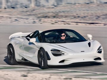 Een witte McLaren 750S sportwagen rijdt over een verharde weg. De auto heeft een strak design met zwarte wielen en een cabriodak.