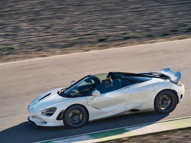 Een witte McLaren 750S cabrioletsportwagen met open dak rijdt op een verharde weg met een onscherpe achtergrond, wat beweging suggereert. De auto heeft een strak, aerodynamisch ontwerp en een zichtbare achtervleugel.