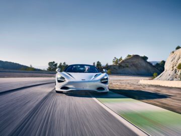 Een witte sportwagen McLaren 750S snelt over een circuit, waarbij een wazige omgeving een hoge snelheid aangeeft. Op de achtergrond zijn ruige heuvels en helderblauwe luchten zichtbaar.