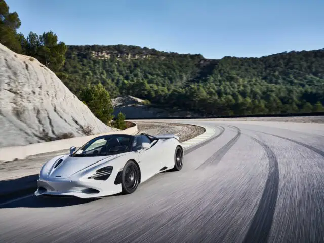 Een McLaren 750S glijdt moeiteloos over een bochtige bergweg omzoomd met bomen en rotswanden onder een helderblauwe lucht.