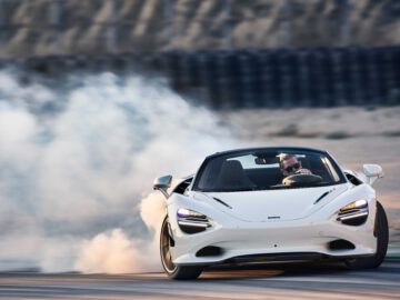 Een witte McLaren 750S zweeft de bocht om op een racecircuit en produceert rook uit de achterbanden. Achter het stuur zit een bestuurder met een zonnebril.