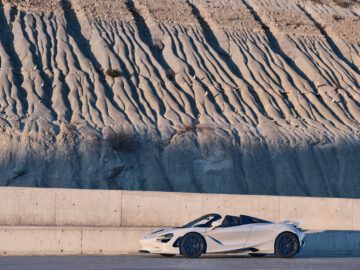 Een witte McLaren 750S staat met open dak geparkeerd op een verhard oppervlak voor een ruige, gelaagde rotsformatie.