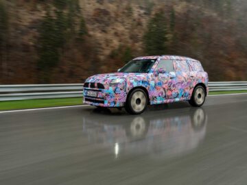 Een gecamoufleerde MINI Countryman met kleurrijke patronen rijdt op een natte, reflecterende weg, grenzend aan een metalen vangrail. Op de achtergrond zijn bomen en een rotsachtige heuvel zichtbaar.