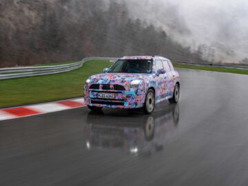 Een MINI Countryman met een kleurrijk ontwerp met patronen rijdt op een natte, mistige weg, navigeert door een bocht terwijl hij reflecteert op het glanzende oppervlak.