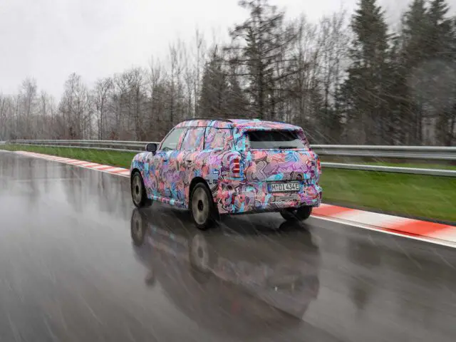 Een gecamoufleerde MINI Countryman rijdt tijdens een regenachtige dag over een natte weg, met bomen op de achtergrond en een slagboom aan één kant.