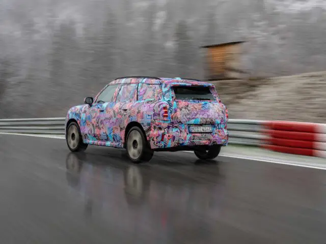 Een gecamoufleerde MINI Countryman SUV rijdt op een nat racecircuit met een onscherpe omgeving, inclusief hekwerk en een constructie op de achtergrond.