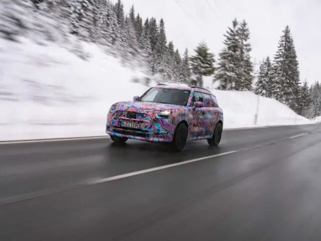 Een gecamoufleerde MINI Countryman SUV rijdt over een met sneeuw omzoomde weg door een bosrijk gebied en toont een kleurrijke, abstracte omhulling terwijl hij met hoge snelheid beweegt.