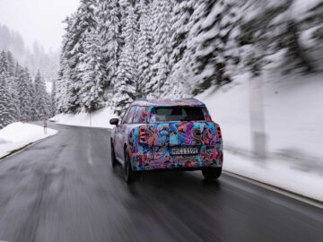 Een MINI Countryman met kleurrijke camouflagekleding rijdt over een besneeuwde, bochtige bergweg omzoomd met met sneeuw bedekte bomen.