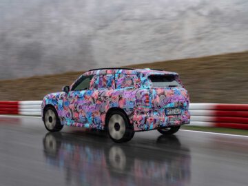 Een gecamoufleerde MINI Countryman rijdt op een natte weg met rood-witte barrières aan de zijkant. Het kleurrijke, abstracte patroon van het voertuig doet vermoeden dat het voertuig wordt getest.