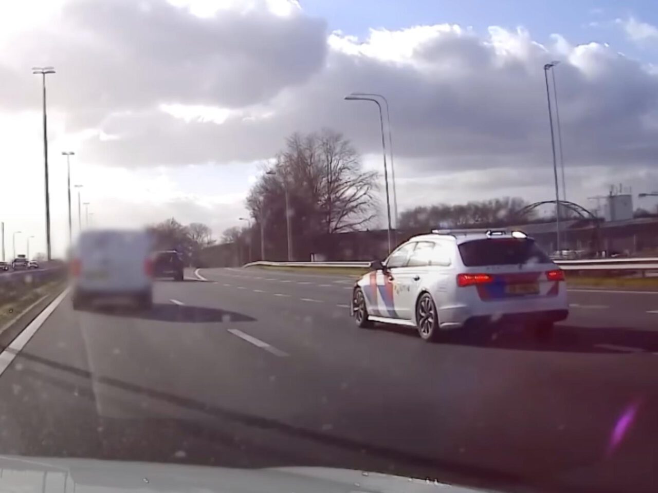Een politieauto rijdt overdag op de rechterrijstrook van een snelweg met meerdere rijstroken, terwijl andere voertuigen verderop zichtbaar zijn. De lucht is gedeeltelijk bewolkt.