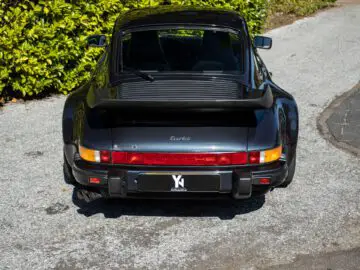 Foto van een zwarte Porsche 911 Turbo, die doet denken aan een klassieke Porsche 930, van achteren gezien, geparkeerd op een grindoprit omringd door groene struiken en bomen.