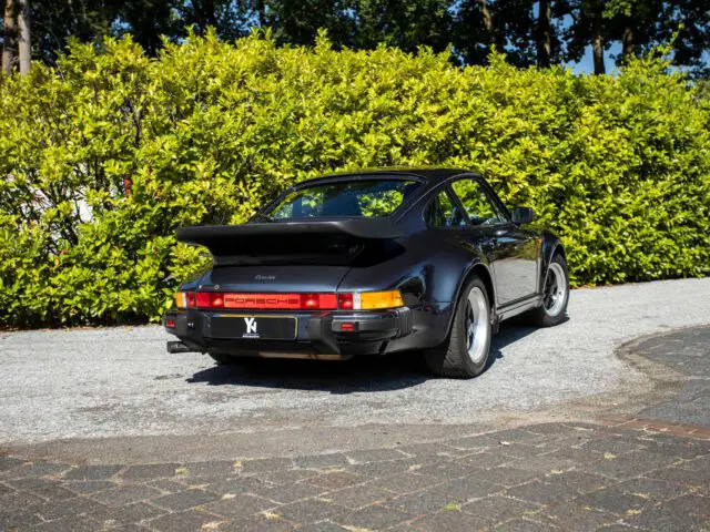 Een zwarte Porsche 911 Turbo, die doet denken aan de iconische Porsche 930, geparkeerd op een verhard terrein met groene struiken op de achtergrond.