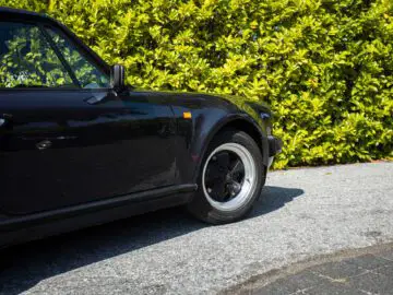 Een zwarte vintage Porsche 930 geparkeerd op een grindoprit met groene struiken op de achtergrond.