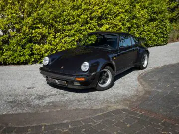 Een strakke zwarte Porsche 911, die lijkt op de iconische Porsche 930, staat geparkeerd op een verharde oprit met grote groene struiken op de achtergrond.