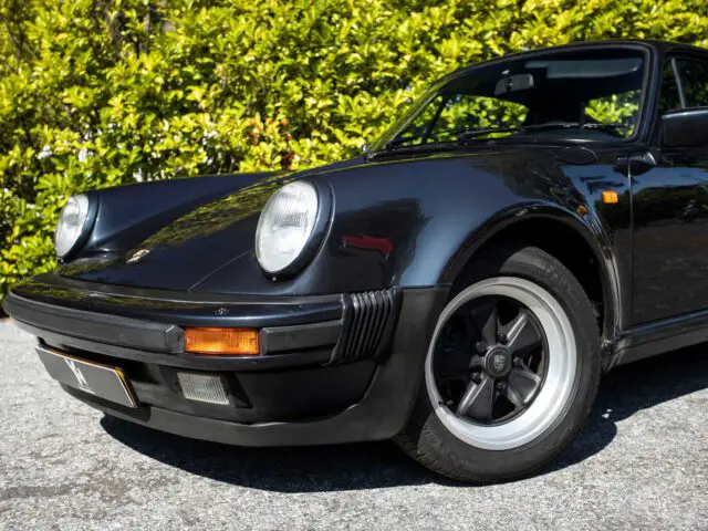 Een zwarte vintage Porsche 930, waarschijnlijk een sportmodel, geparkeerd op een gecementeerde grond met groene struiken op de achtergrond.