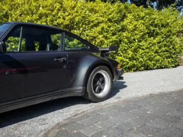 Op een verharde oprit met een heg op de achtergrond staat een zwarte Porsche 930 sportwagen geparkeerd. Alleen de achterkant van de auto is zichtbaar.