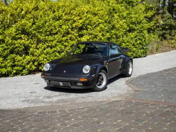 Een zwarte Porsche 930 klassieke sportwagen staat geparkeerd op een oprit met weelderig groen op de achtergrond.