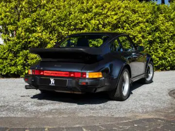 Een zwarte klassieke Porsche 930 sportwagen staat geparkeerd op een onverharde oprit, met weelderig groen op de achtergrond. De auto beschikt over een opvallende achterspoiler en een 'Turbo'-badge op de achterkant.