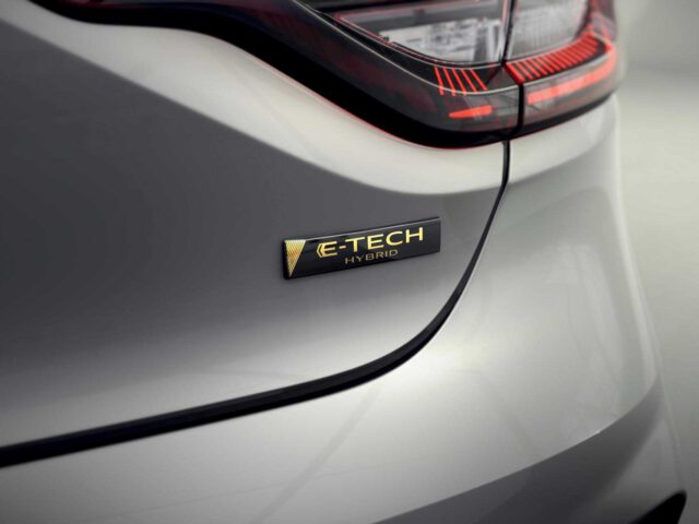 Close-up van het achterembleem van een hybride auto van Renault Clio met de tekst "E-TECH Hybrid" op een donker rechthoekig plaatje met metalen letters. Het achterlicht en een deel van de zilveren achterkant van de auto zijn zichtbaar.