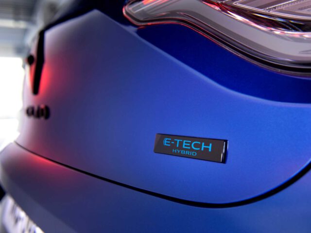 Close-up van de achterkant van een blauwe Renault Clio met het "E-Tech Hybrid"-embleem en een deel van een achterlicht. De achtergrond is wazig.