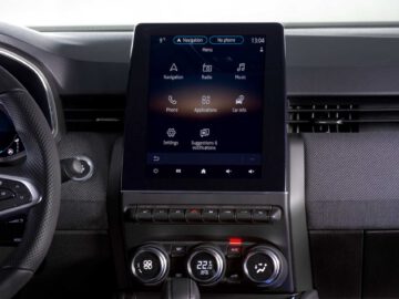 Close-up van de middenconsole van een Renault Clio met een groot touchscreen waarop navigatie en verschillende applicaties worden weergegeven, met daaronder bedieningsknoppen en klimaatknoppen.