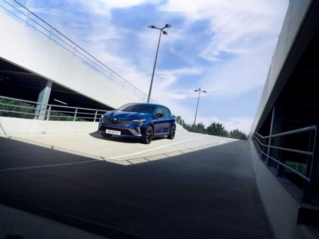 Op een heldere dag staat een blauwe Renault Clio geparkeerd op een helling in een parkeergarage met meerdere verdiepingen, met straatverlichting en balustrades zichtbaar op de achtergrond.