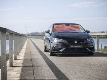 Een zwarte SEAT Leon Cupra 300 staat geparkeerd op een brug over een watermassa. De lucht is helder met enkele wolken en er is een windturbine zichtbaar op de achtergrond.