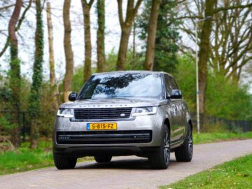 Een zilveren Range Rover SUV rijdt op een smal, met bomen omzoomd pad met een kentekenplaat met de tekst "S-815-FZ".