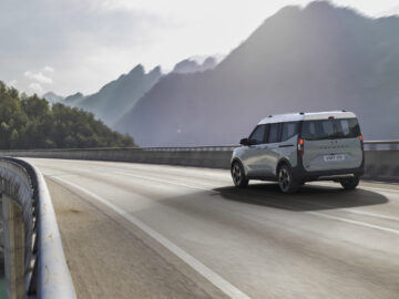 Een Ford E-Tourneo Courier rijdt op een verhoogde snelweg met bergen en bomen op de achtergrond onder een bewolkte hemel.