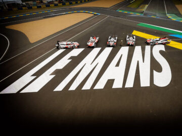 Vijf sportwagens, waaronder een strakke Toyota, staan opgesteld op het circuit van Le Mans, met de grote witte tekst "LE MANS" op het baanoppervlak geschilderd.