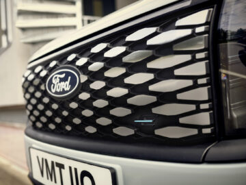 Close-up van de grille van een Ford E-Tourneo Courier met een zeshoekig patroon en het Ford-logo in het midden met daaronder een zichtbare kentekenplaat.