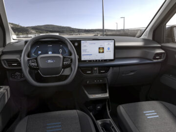 Binnenaanzicht van een Ford E-Tourneo Courier met een Ford-logo op het stuur, een digitaal dashboard en een groot centraal touchscreen-display, met twee stoelen op de voorgrond.