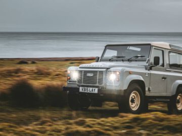 Een zilveren Classic Defender met kenteken "V815 LAY" rijdt in de schemering over een grasachtig terrein vlakbij de zee.