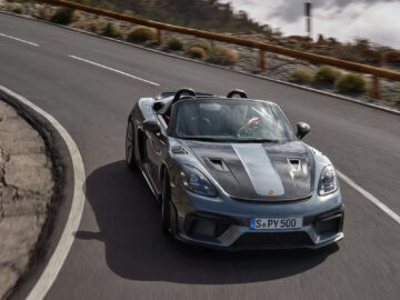 Een zwart-grijze Porsche 718 Spyder RS rijdt over een bochtige bergweg, omlijst door een bewolkte lucht op de achtergrond.