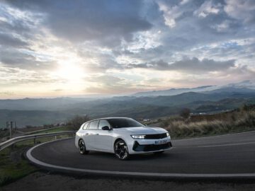 Een witte Opel Astra Sports Tourer GSe, met 225 pk, rijdt bij zonsondergang over een bochtige bergweg, met glooiende heuvels en een bewolkte lucht op de achtergrond.