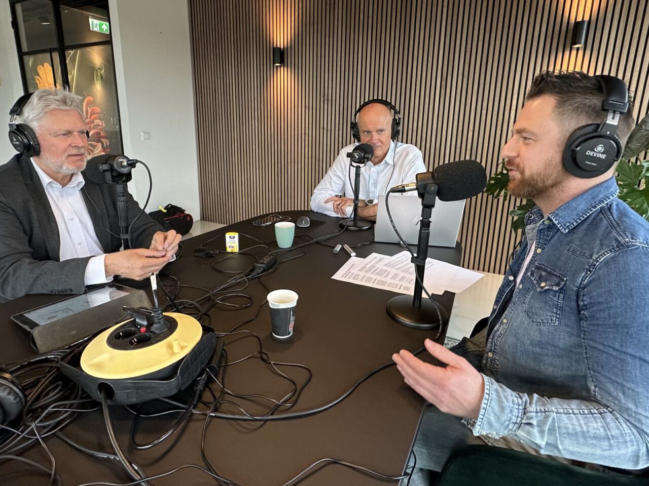 Aan een tafel met microfoons zitten drie mannen met koptelefoon op en zijn in gesprek over verkeersdoden. Op tafel liggen aantekeningen, een laptop en een koffiekopje. De achtergrond bestaat uit verticale houten latten en planten.