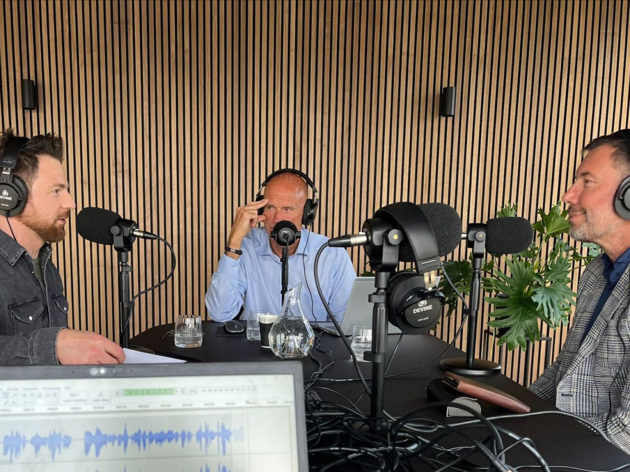 Drie mannen nemen de AutoRAI Podcast op rond een tafel met microfoons en koptelefoons; op de voorgrond staat een laptop die audiogolfvormen weergeeft.